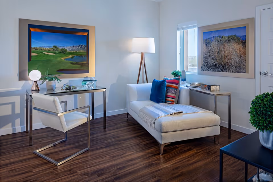A modern interior room with a white chaise lounge adorned with colorful pillows and a throw blanket. Next to it is a small metal side table with books and decorative items. A floor lamp with a white shade stands behind the chaise. Across from it is a sleek glass desk with a white cushioned chair, a small lamp, a plant, and some books. The room has wooden flooring and white walls decorated with two large framed landscape photographs. A window lets in natural light.