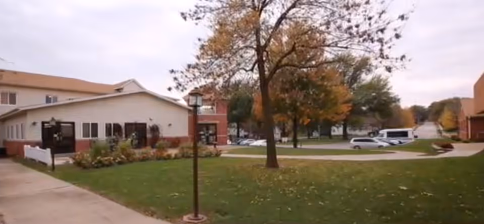 Front exterior of a senior living community with a lawn, trees, walkways, and parked vehicles near the building.