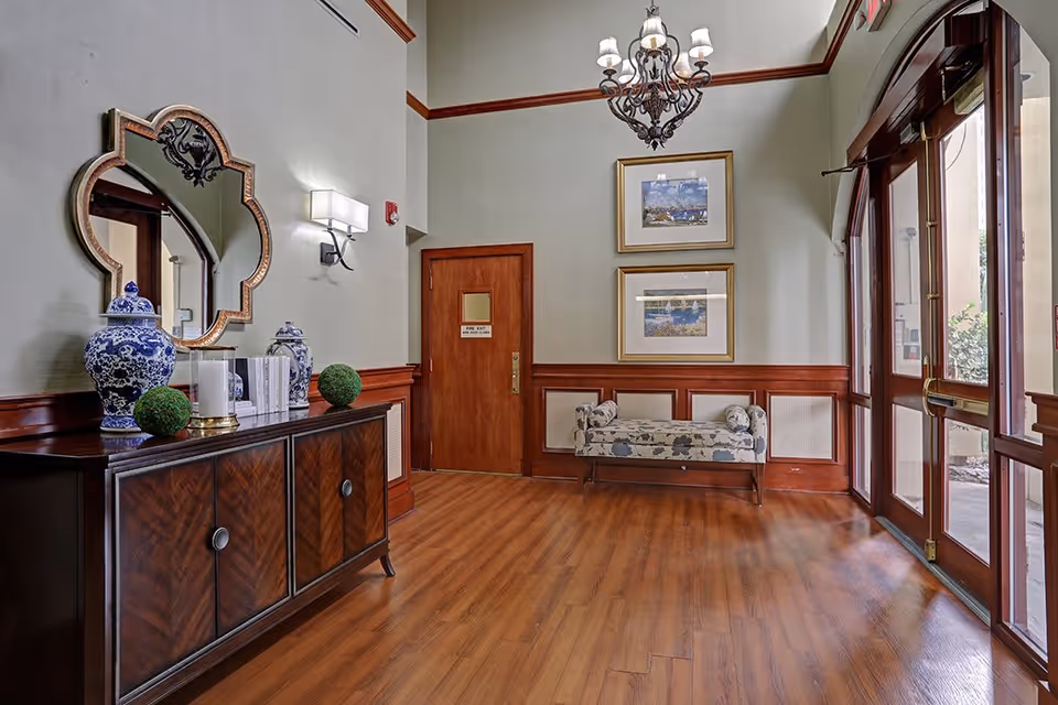 An elegant interior entryway with wooden flooring and wood-paneled walls. A decorative mirror and blue-and-white vases sit on a wooden cabinet on the left. A floral upholstered bench is placed against the far wall beneath two framed pictures. A chandelier hangs from the ceiling, and glass double doors on the right lead outside.