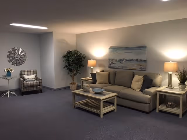 A cozy living room area with a beige sofa adorned with multiple cushions, two wooden side tables each with a lamp, a wooden coffee table with decorative items, a potted plant in the corner, a plaid armchair with a pillow, a small round table with a vase of sunflowers, and a wall decoration resembling a clock and a painting of a beach scene on the wall.