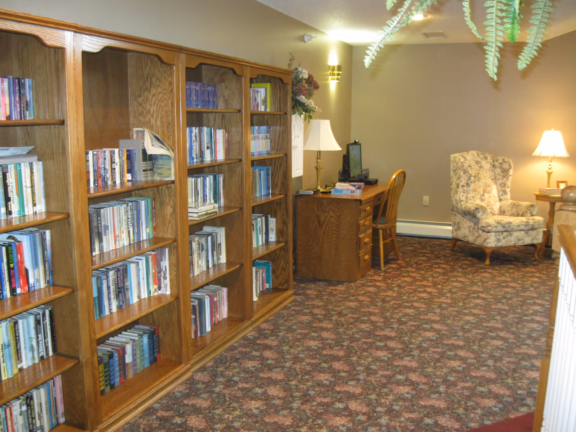 Carpeted common room with wooden bookshelves, a desk with a computer and lamp, and a floral upholstered armchair.