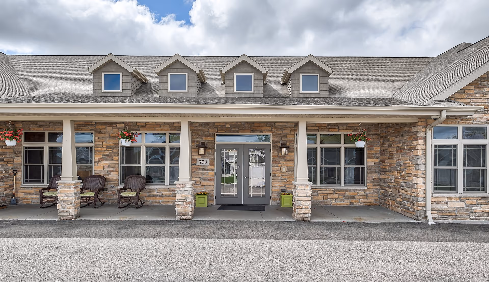 Front exterior view of a single-story building with stone facade, multiple windows, and a covered porch with columns. There are hanging flower pots and wicker chairs on the porch. The building has a gray shingled roof with four small dormer windows and double glass entrance doors.