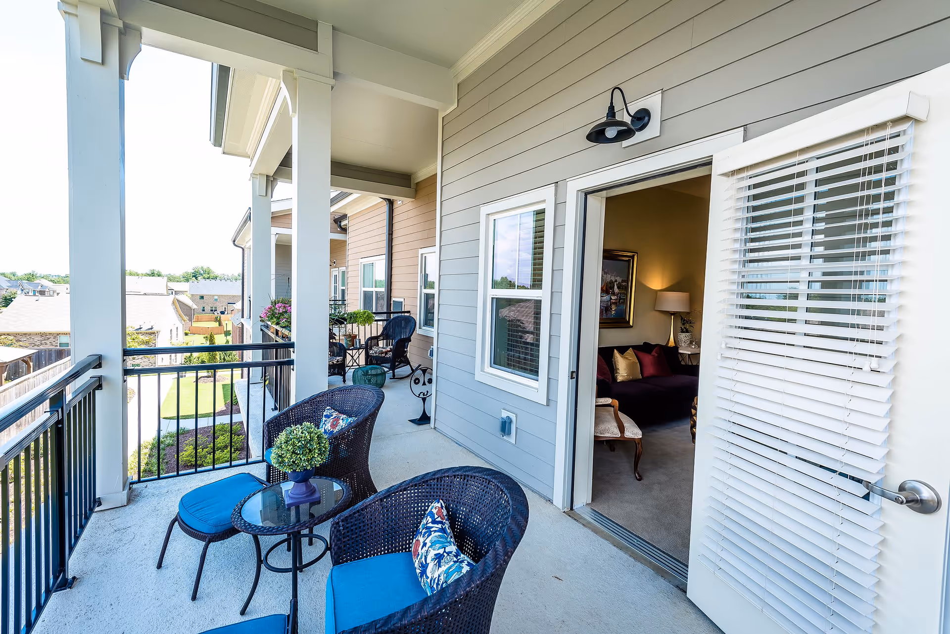 A covered balcony with two wicker chairs and matching ottomans with blue cushions, a small round glass table with a potted plant, and a view of neighboring buildings and greenery. The balcony has white pillars and black metal railings. A door with white blinds is open, leading into a living room with a dark sofa, a lamp, and framed artwork on the wall.