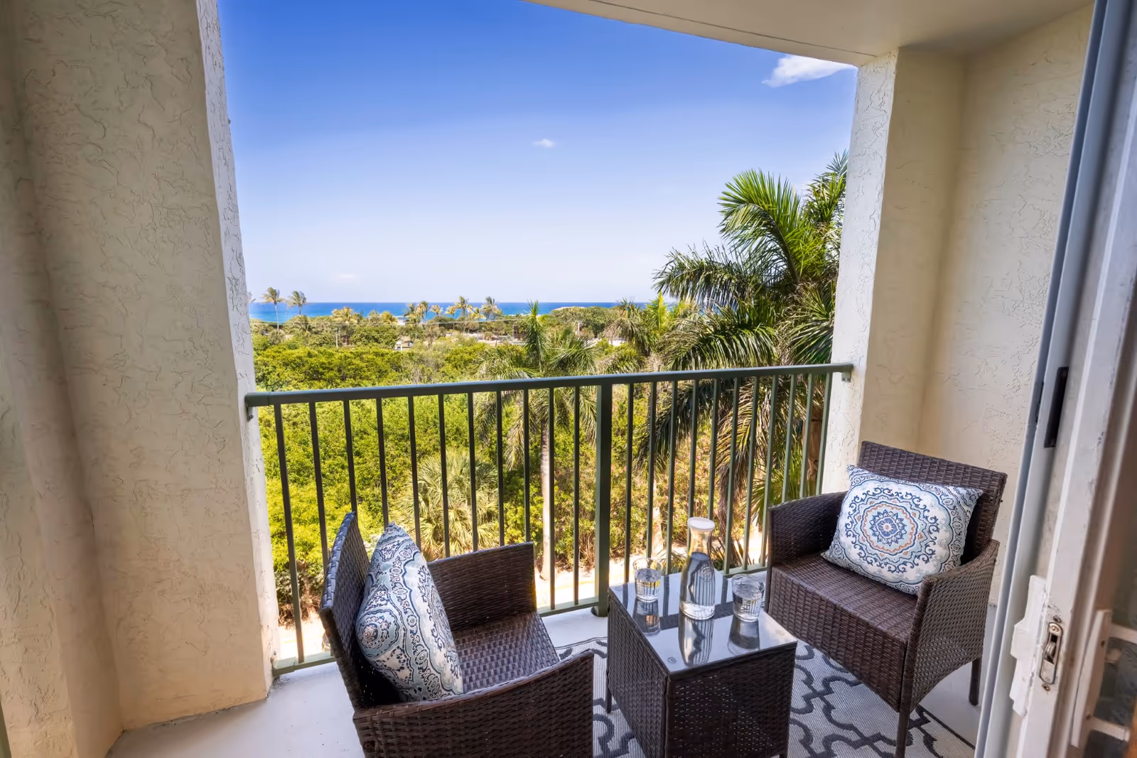 Small furnished balcony with two wicker chairs, a table with glassware, and decorative pillows overlooking palm trees and the ocean.