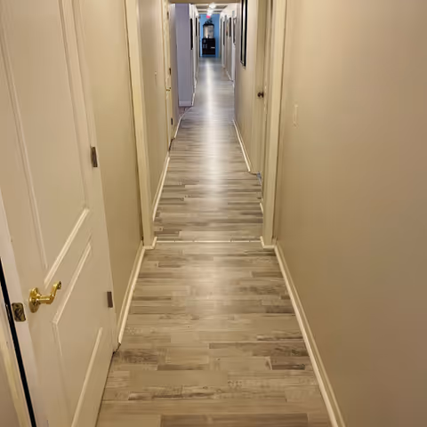 A long, narrow hallway with light-colored wooden flooring and beige walls. Several closed doors line both sides of the hallway, and framed pictures hang on the right wall. The hallway is well-lit with ceiling lights and extends into the distance.