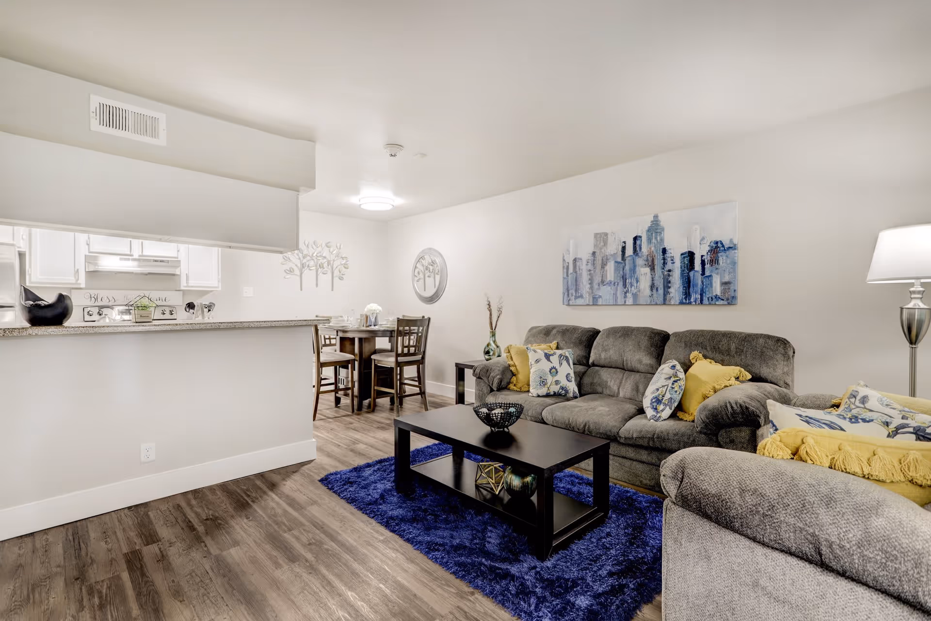 Spacious living room with gray sofas, a black coffee table on a blue rug, and a view into the kitchen and dining area.