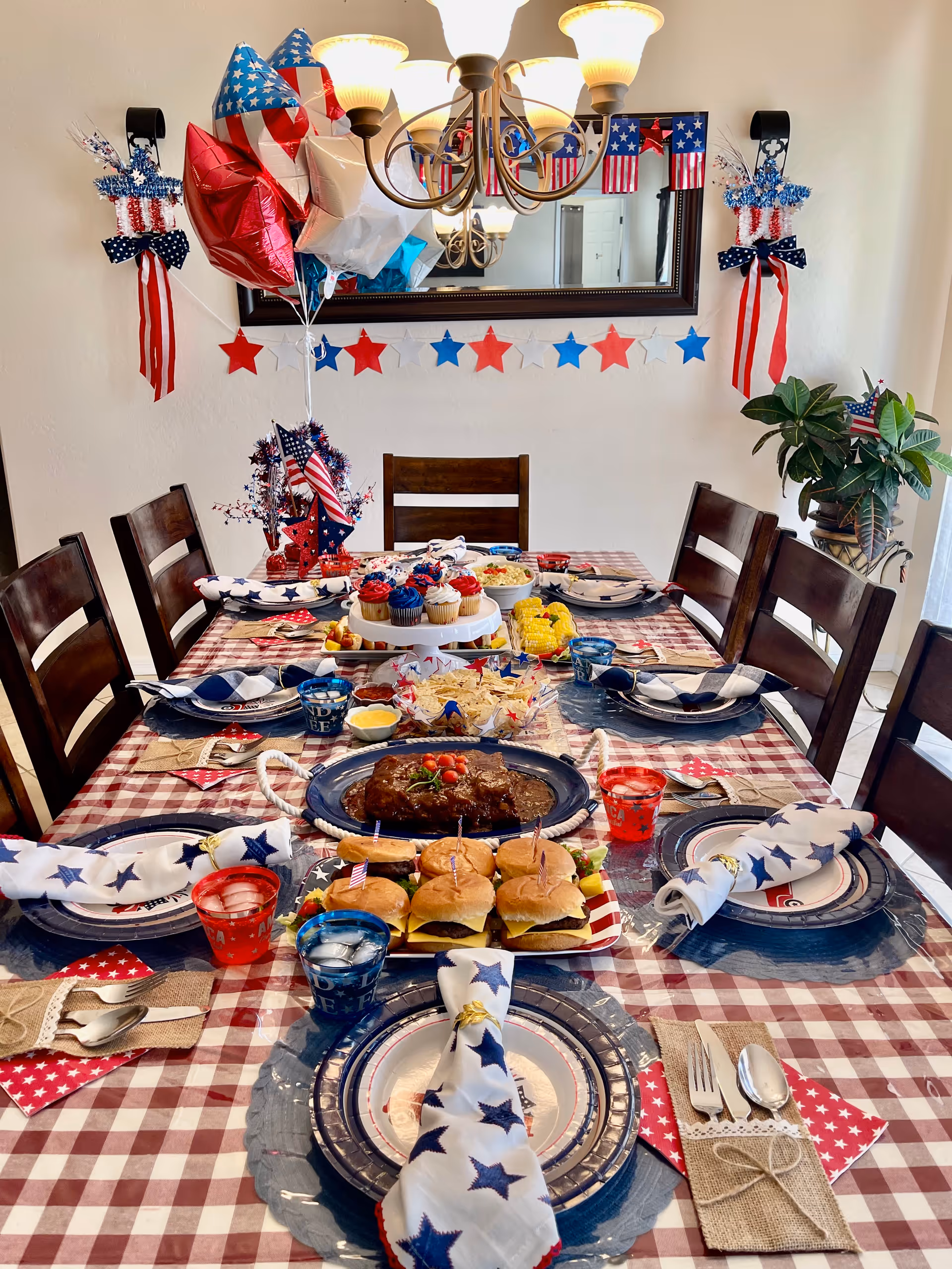 A dining table set for a patriotic-themed meal with red, white, and blue decorations including star-patterned napkins, American flag balloons, and star garlands. The table has plates, utensils, and glasses filled with water and red drinks. Food on the table includes sliders, a meatloaf, corn on the cob, cupcakes, and chips with dip. The room has wooden chairs, a large mirror on the wall, and a chandelier overhead.