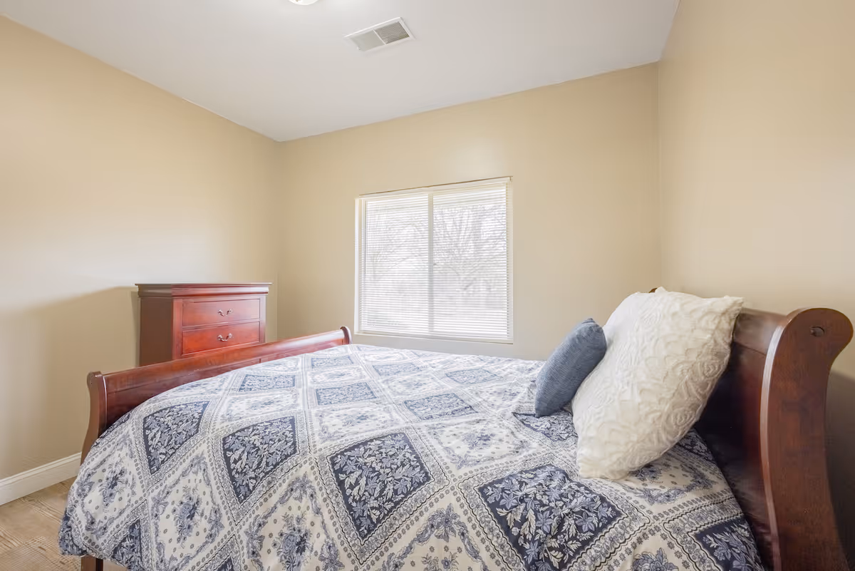 A bright bedroom with beige walls featuring a wooden bed with a blue and white patterned bedspread and two pillows, one white and one blue. There is a wooden dresser with three drawers against the wall and a window with blinds letting in natural light.