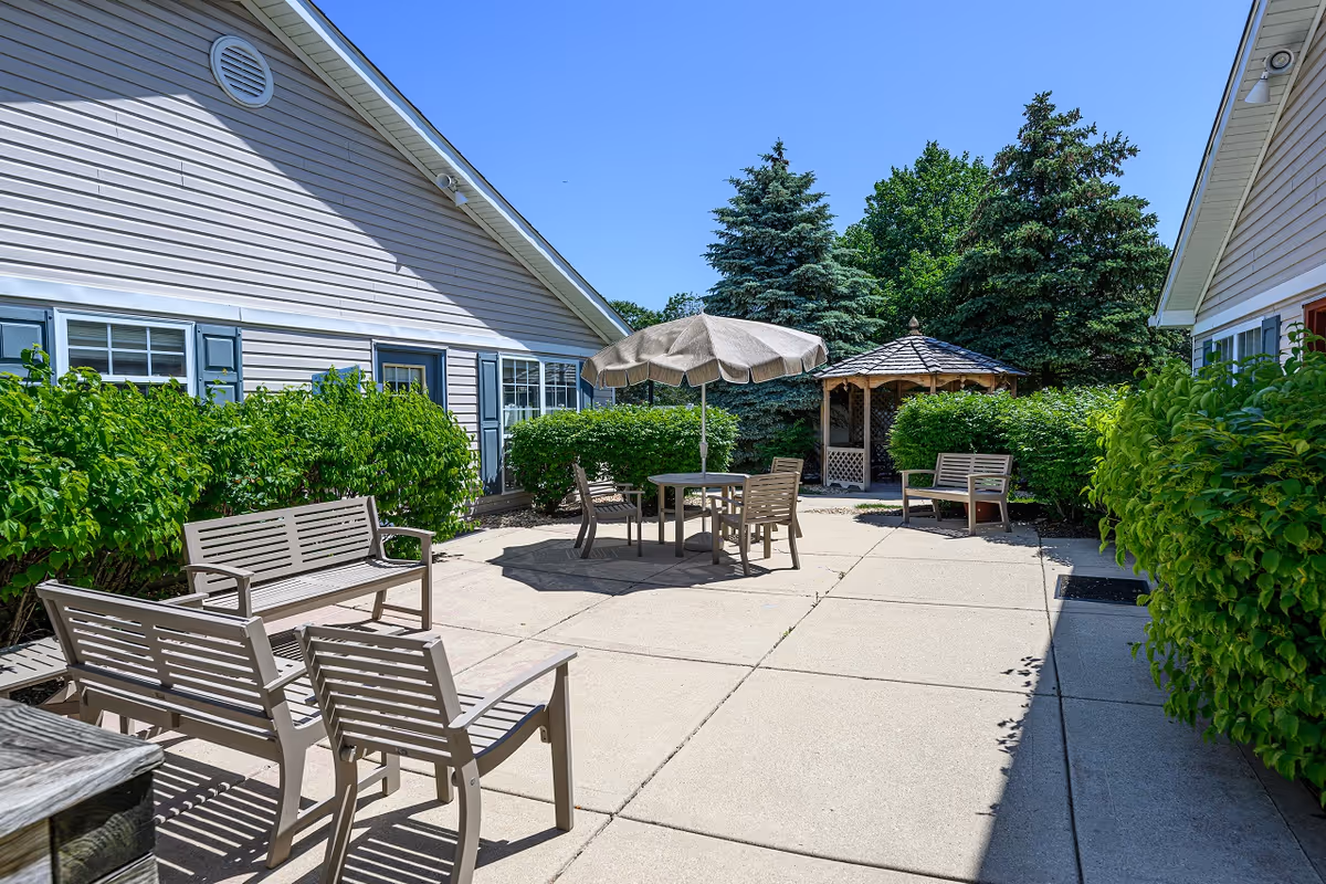 Outdoor patio area at Arden Courts of Geneva featuring several benches and chairs arranged around a table with a large umbrella. The patio is surrounded by green bushes and trees, with a small gazebo visible in the background. The sky is clear and blue.