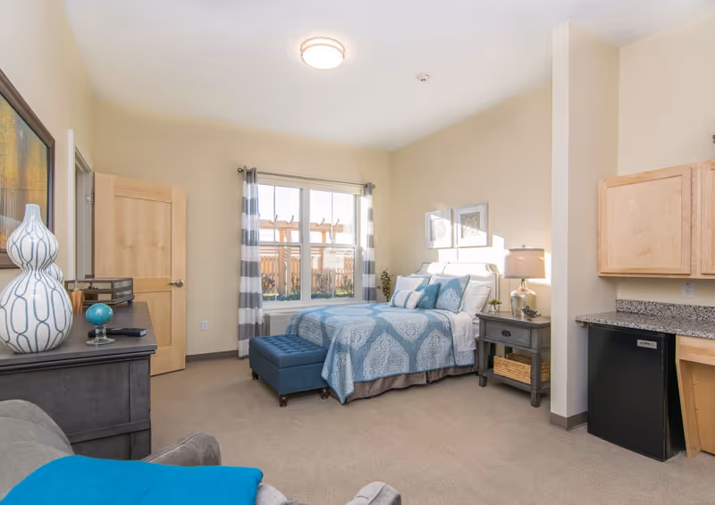 Bright furnished studio-style bedroom with a bed, nightstand, dresser, window, and a small kitchenette area.