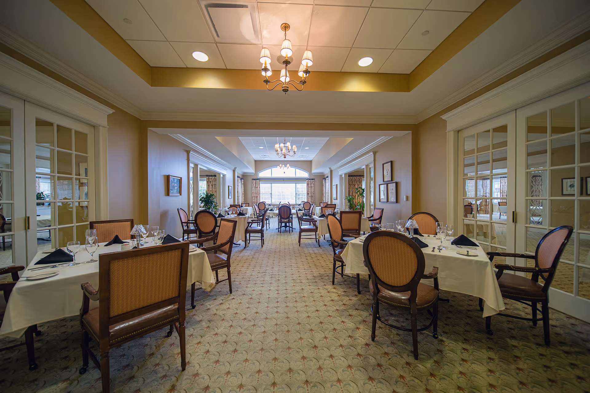 Spacious formal dining room with tables set with white linens and place settings, upholstered chairs, chandeliers, and large windows at the far end.