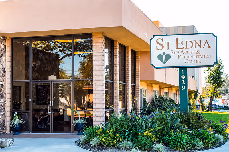 Exterior front view of St. Edna Sub-Acute and Rehabilitation Center building with large windows, a glass entrance door, and a sign displaying the facility name and address number 1929. The area in front of the building features green plants and landscaping.