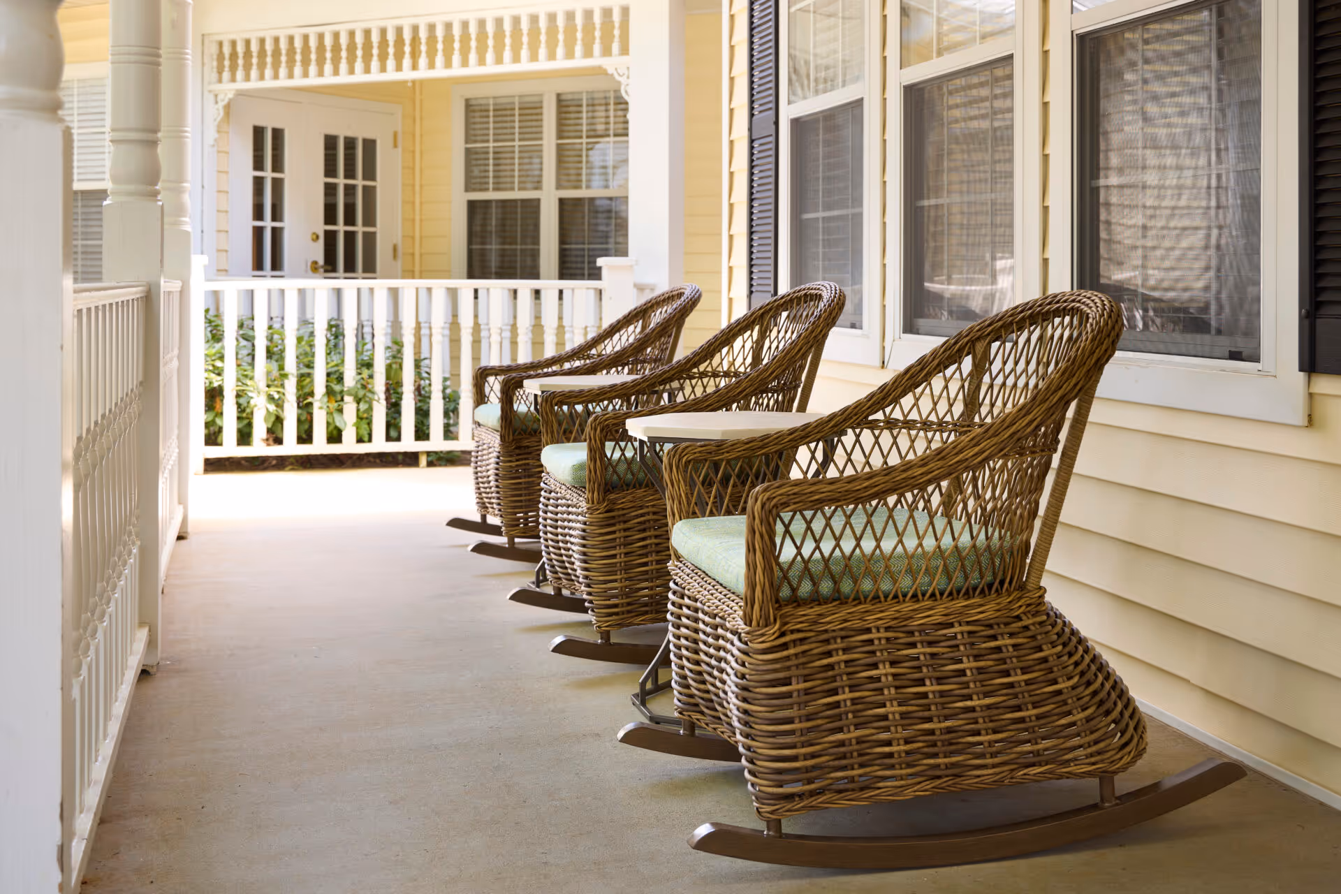 A porch area with three wicker rocking chairs with green cushions lined up against the yellow siding of a building. The porch has white railings and columns, and there are windows with black shutters on the building wall.