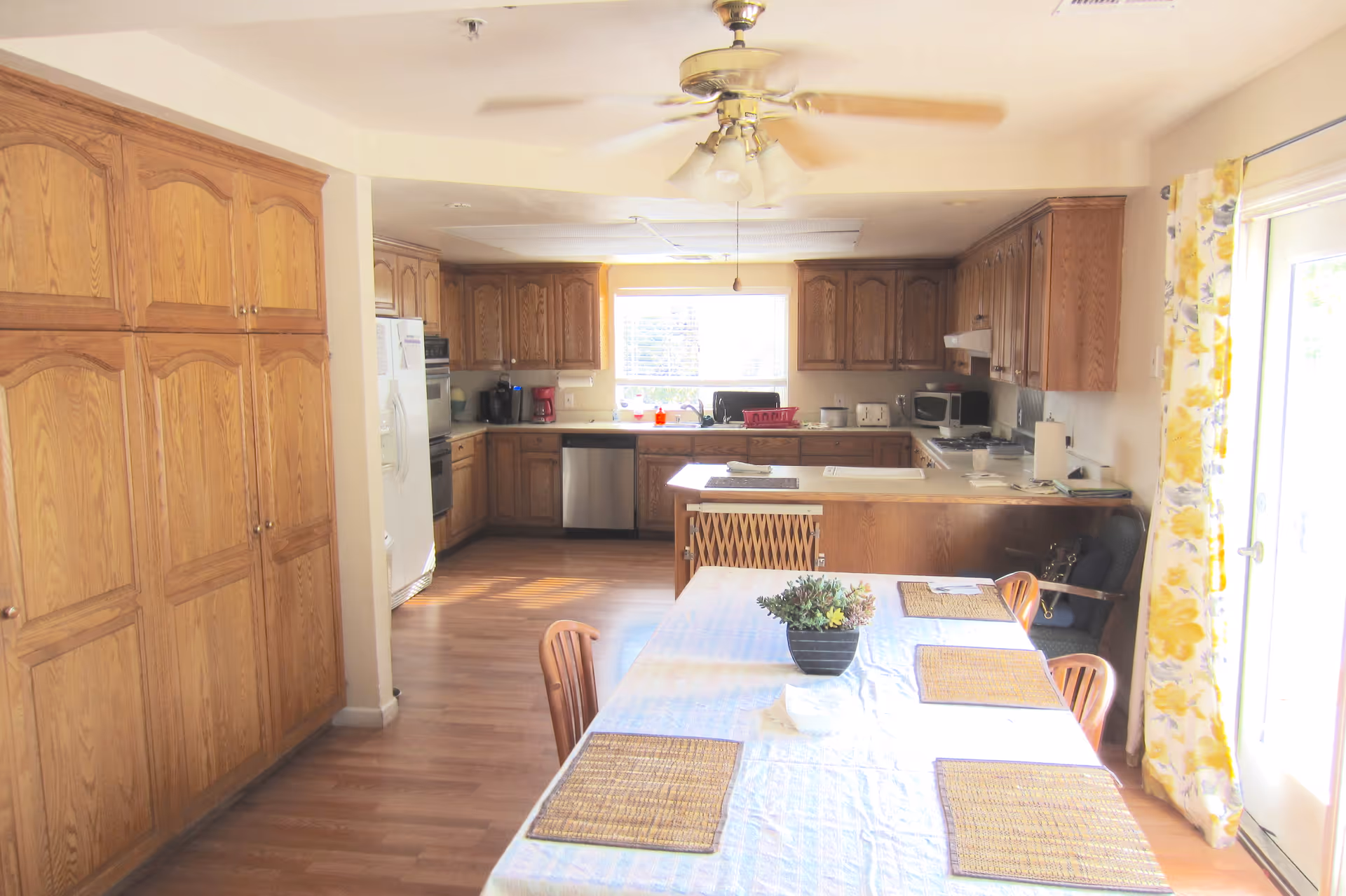 Bright kitchen and dining area with wooden cabinets, a ceiling fan, and a table set with placemats and a small plant centerpiece. Sunlight streams in through a window and glass door with yellow floral curtains.