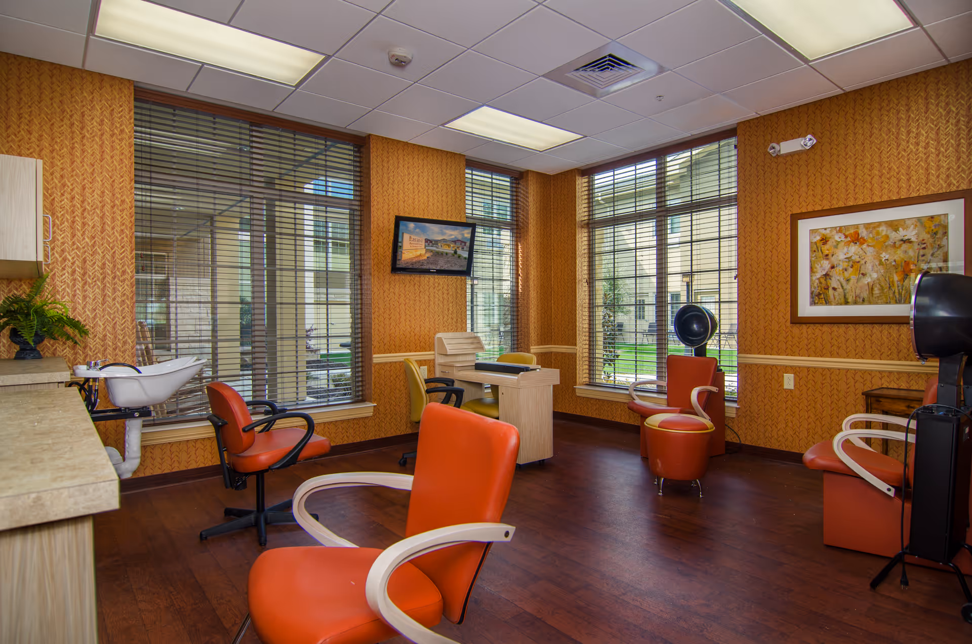 Interior view of a salon area in a senior living facility with orange salon chairs, a hair washing station, a hair dryer, large windows with blinds, a desk with chairs, and framed artwork on the wall.