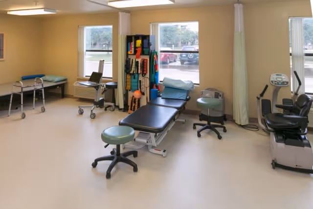 A physical therapy room with exercise equipment including a treatment table, stools, a walker, resistance bands, and a stationary recumbent bike. The room has large windows letting in natural light and light-colored walls and flooring.