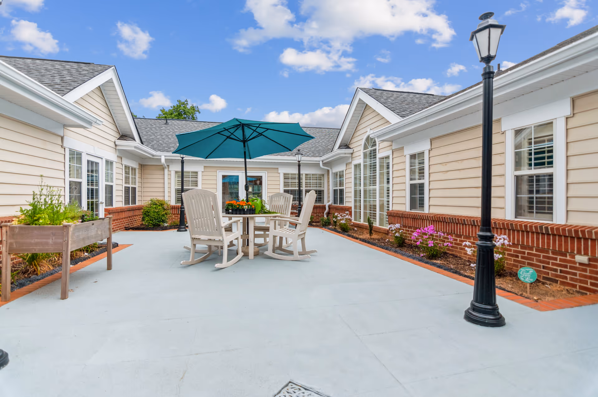 Outdoor courtyard area at Legacy Heights Senior Living Community featuring a round table with four white chairs and a large green umbrella. The courtyard is surrounded by beige buildings with white trim and brick accents. There are flower beds with plants and flowers along the edges, and two black lamp posts are visible. The sky is blue with scattered clouds.