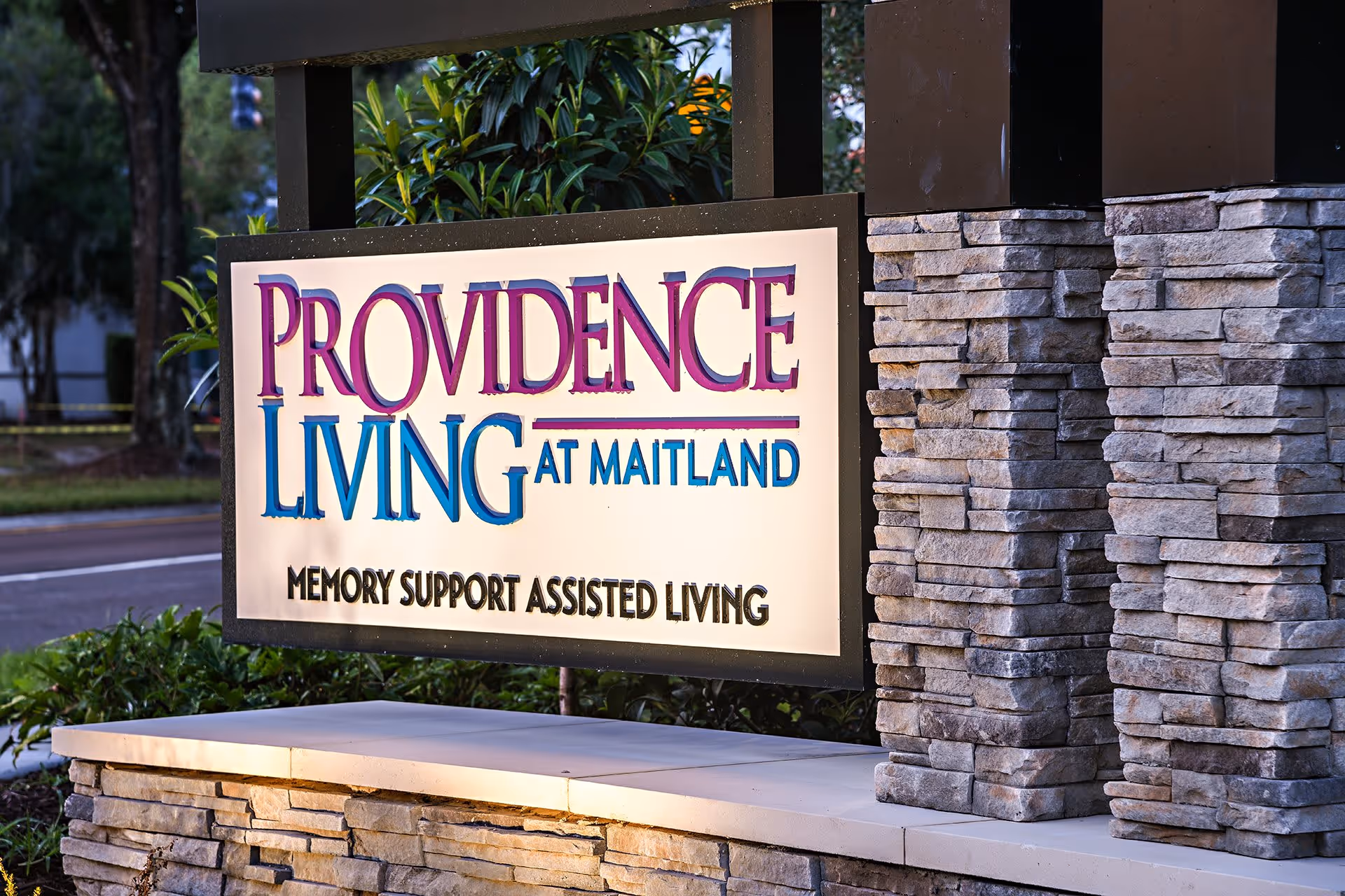 Outdoor sign for Providence Living at Maitland, a memory support assisted living facility, mounted on a stone base with greenery and a road in the background.