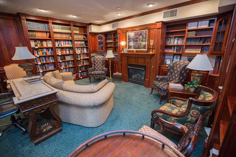 A cozy library room with wooden bookshelves filled with books lining the walls. The room features a fireplace with a painting above it, several upholstered armchairs, a curved sofa, a wooden desk with a lamp, and a green carpeted floor. The lighting is warm with recessed ceiling lights and table lamps.