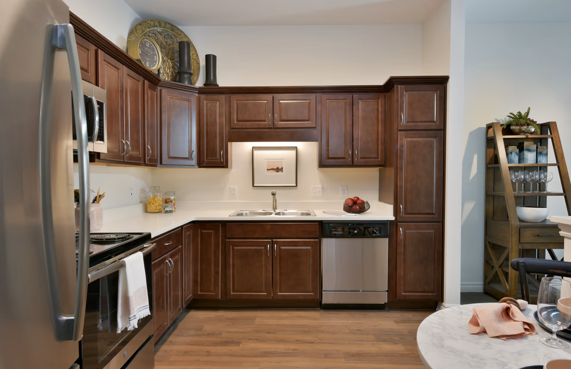 Modern kitchen with dark wood cabinets, stainless steel refrigerator, oven, microwave, and dishwasher. White countertops with a double sink and a framed picture on the wall above the sink. A wooden shelving unit with glassware and decorative items is visible in the background. A round table with a pink napkin and glassware is partially visible in the foreground.