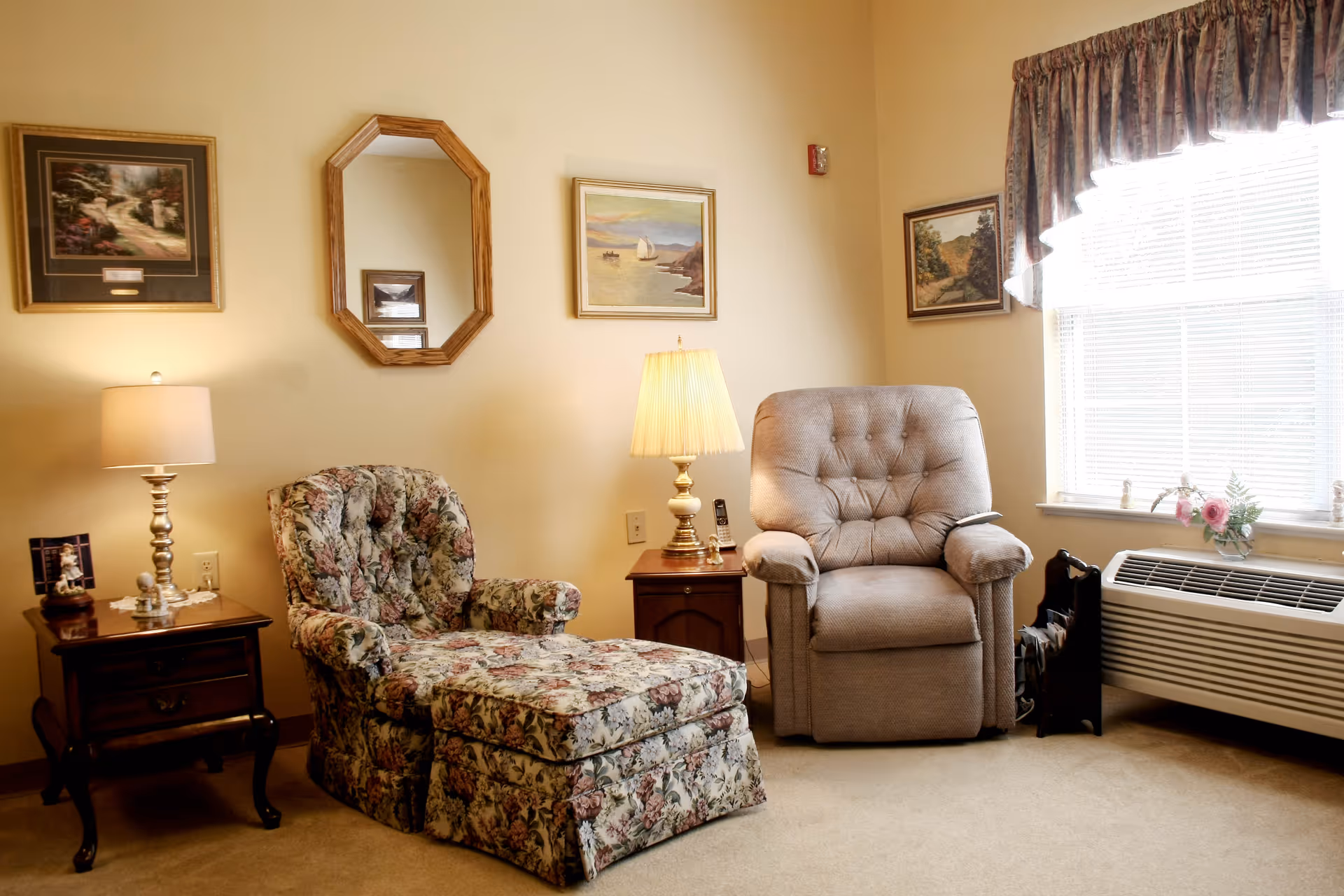 A cozy sitting area in a senior living facility featuring a floral upholstered armchair with an ottoman, a beige recliner, two wooden side tables each with a lamp, framed paintings and a mirror on the wall, a window with curtains, and a heating/cooling unit below the window.