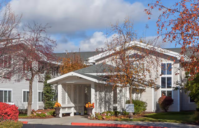 Exterior view of a senior living facility named Trustwell Living at Ridgeview Place, showing a light-colored building with a covered entrance, surrounded by trees with autumn foliage and landscaped bushes under a partly cloudy sky.