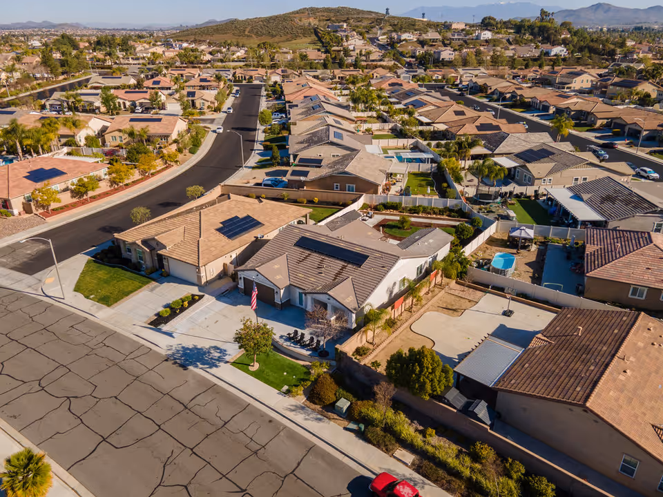 Aerial view of a suburban neighborhood with single-story houses, streets, and some greenery. The houses have tiled roofs, some with solar panels, and fenced backyards. The area is surrounded by hills and mountains in the distance under a clear sky.