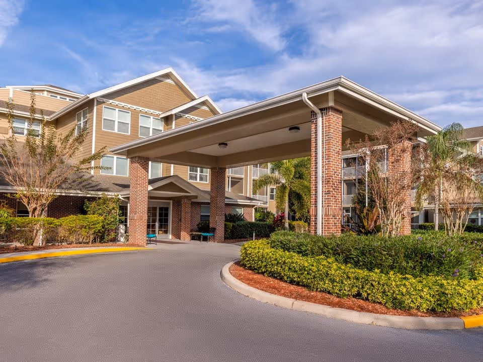 Exterior view of Desoto Beach Club - Sky Active Living facility showing a covered entrance with brick pillars, surrounded by landscaped greenery and a clear sky above.
