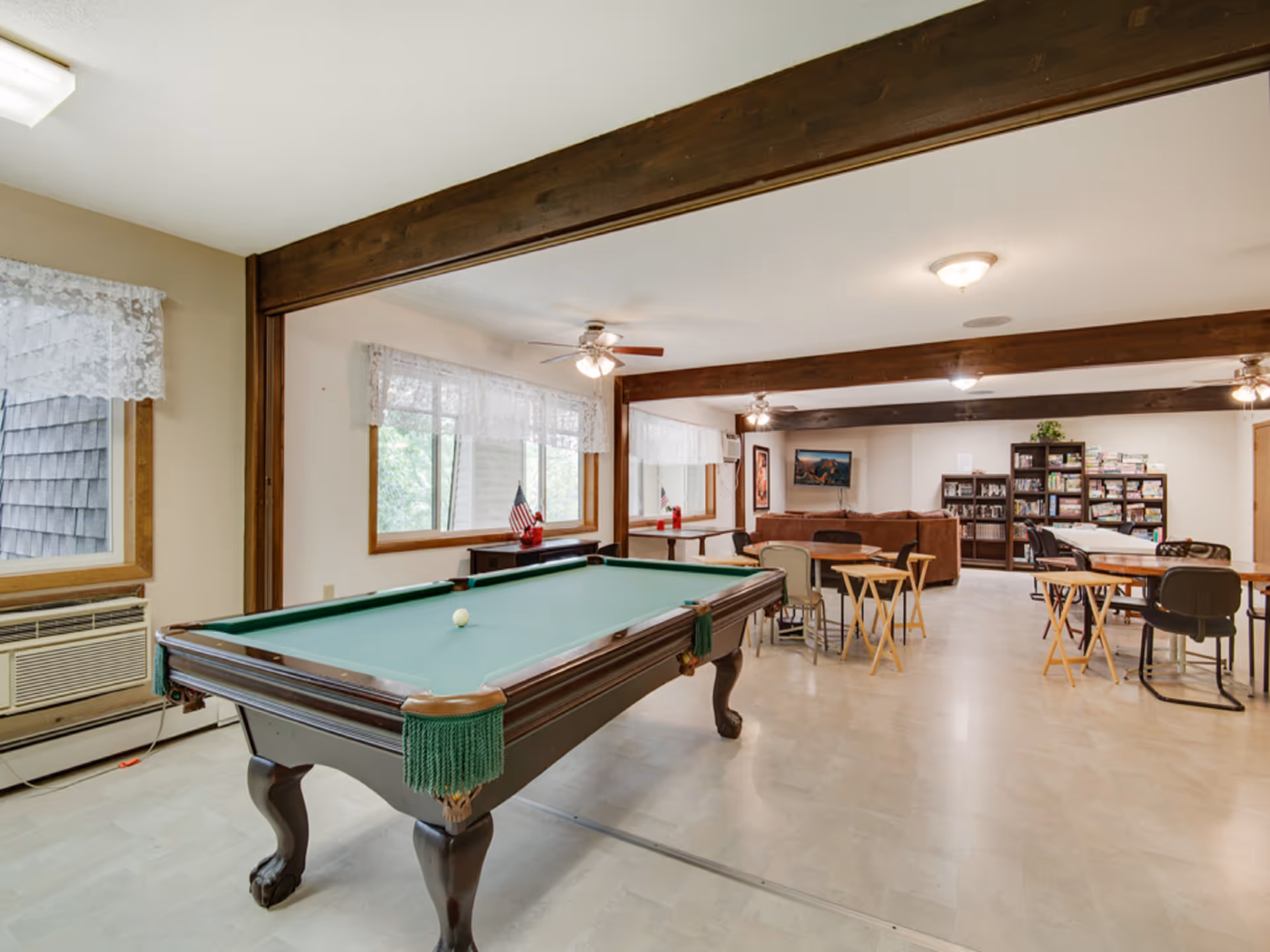 A spacious recreational room with a pool table in the foreground, several tables and chairs arranged for games or activities, a brown sectional sofa, bookshelves filled with books and games, and large windows with lace curtains letting in natural light. The room features wooden beams on the ceiling and ceiling fans.