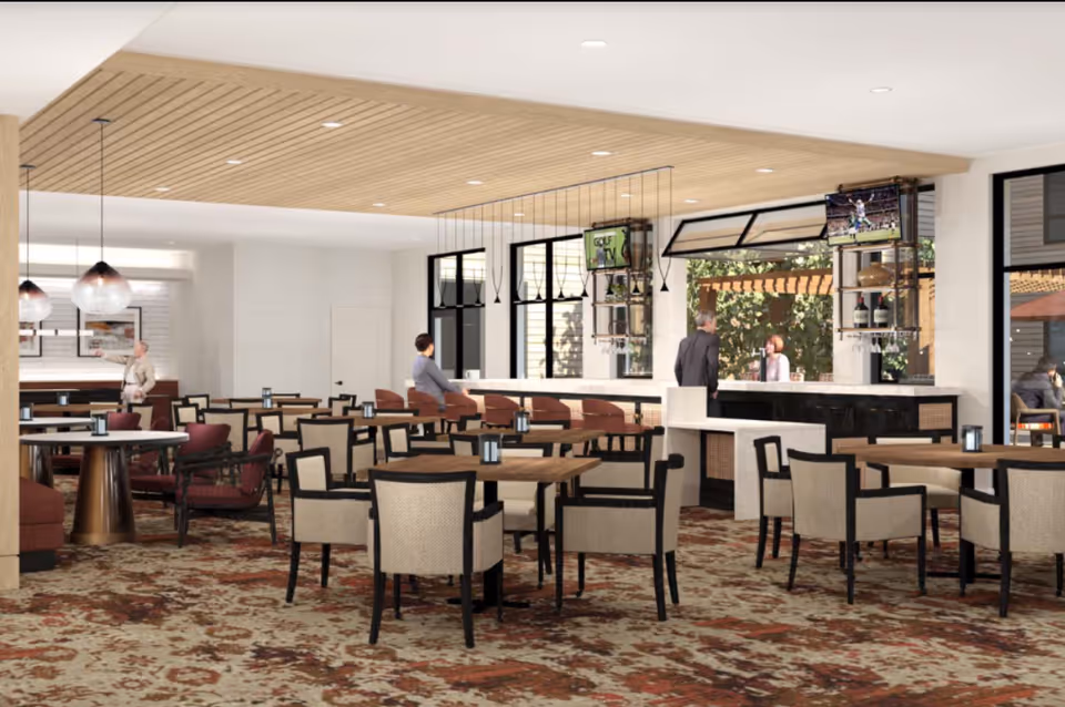 A spacious dining area in a senior living facility featuring multiple tables with chairs, a bar with stools, pendant lighting, and large windows. Several people are seen seated and standing, with a patterned carpet and a wooden slatted ceiling adding warmth to the space.
