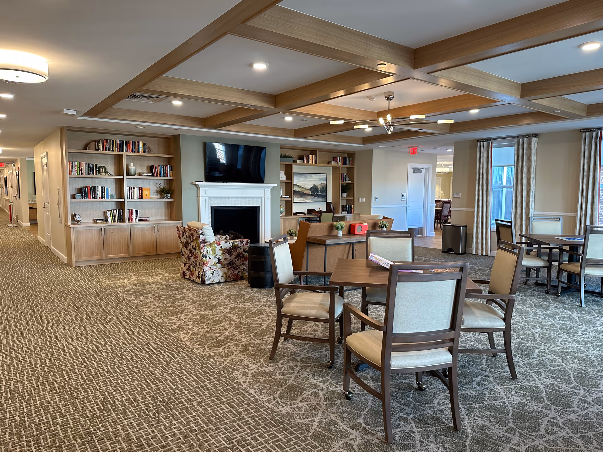 A spacious and well-lit common area in a senior living facility featuring a patterned carpet, several tables with chairs, a cozy seating area with a floral armchair and a brown sofa, built-in wooden bookshelves filled with books, a white fireplace with a mounted flat-screen TV above it, and large windows with curtains allowing natural light to enter.