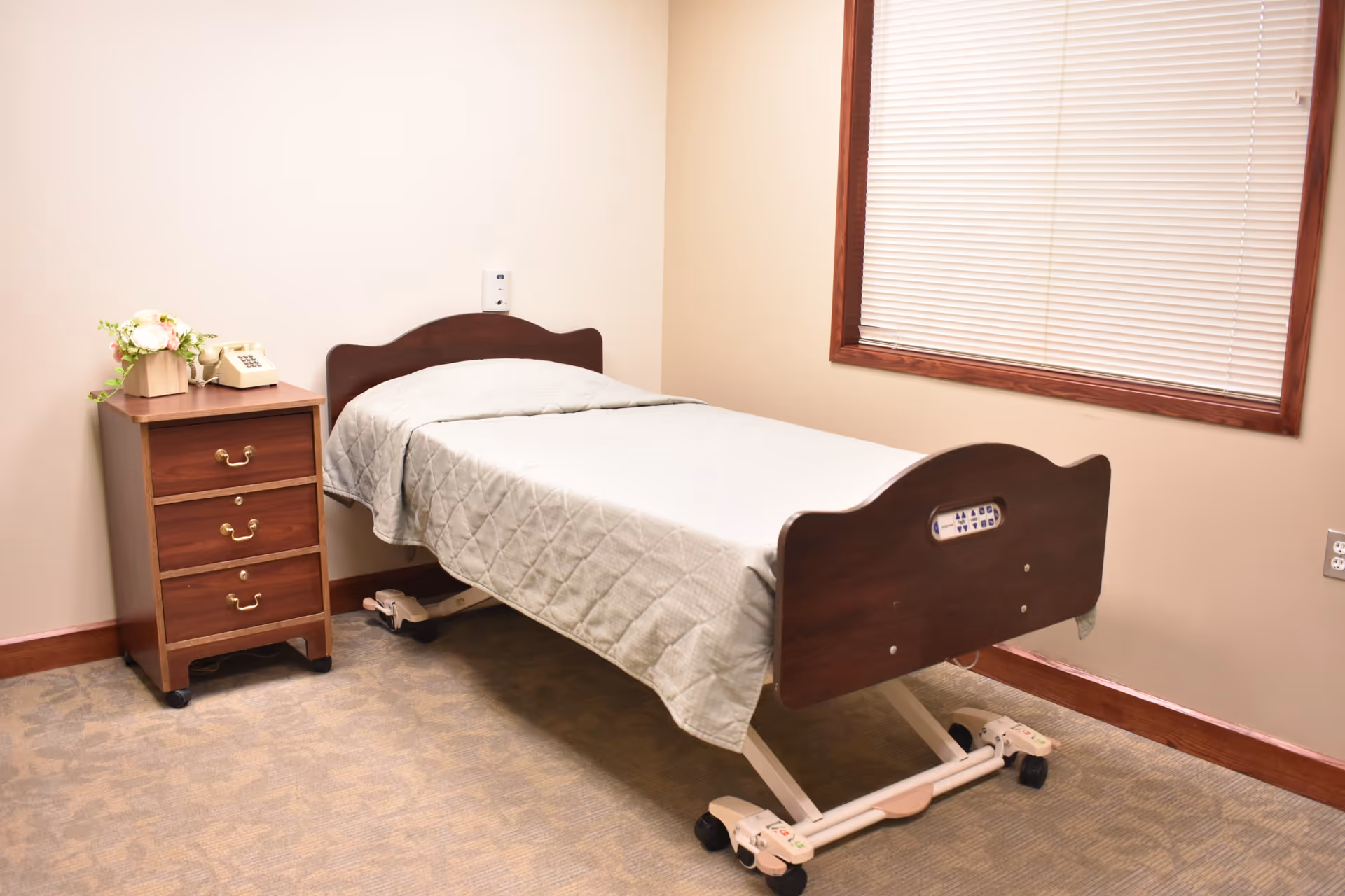 A simple senior care bedroom with an adjustable hospital bed, wooden nightstand holding a phone and flowers, and a window with closed blinds.