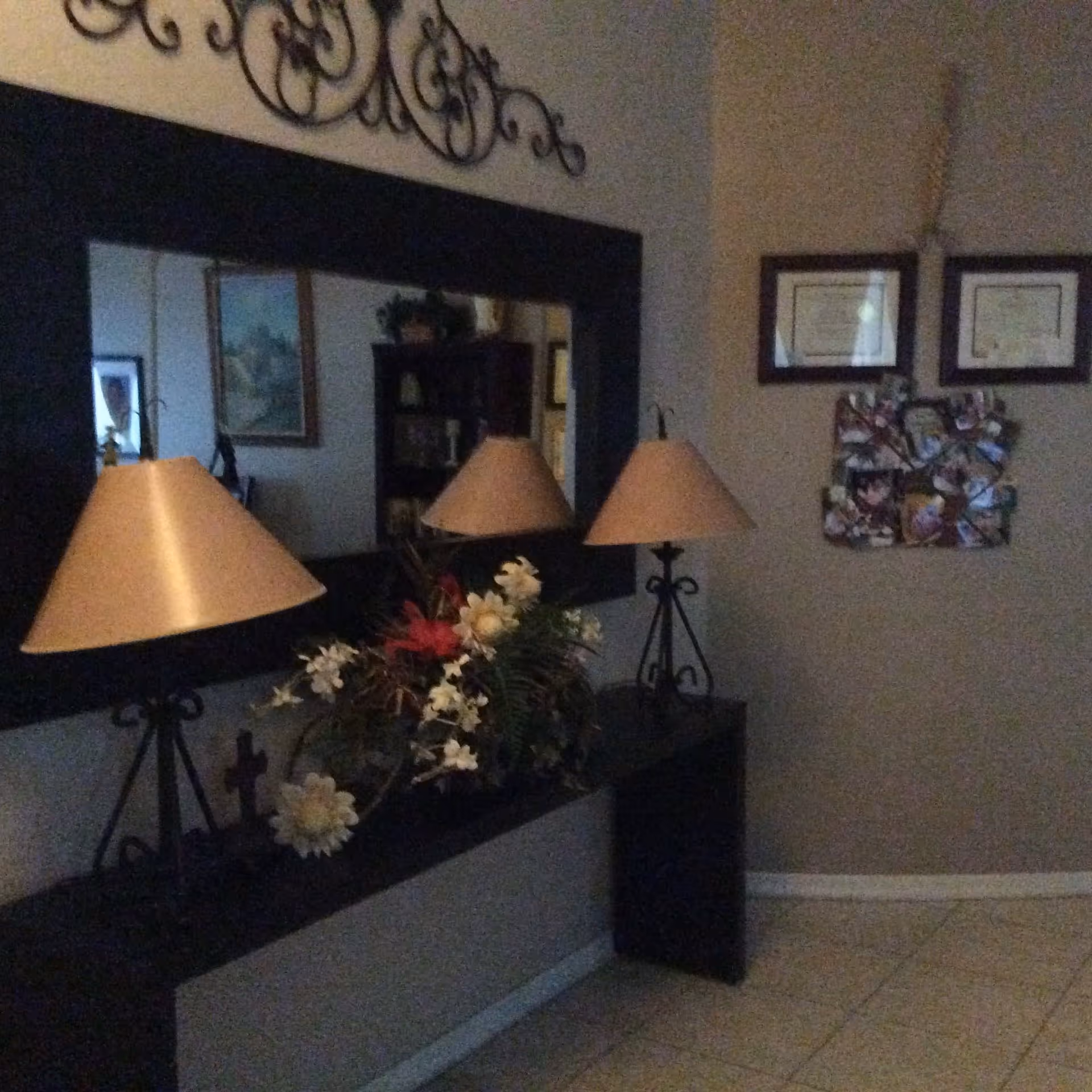 Interior corner of a room with a large rectangular mirror above a black console table. On the table are two lamps with beige shades and a floral arrangement. The wall above the mirror has decorative wrought iron art. On the adjacent wall, there are two framed certificates and a photo collage.