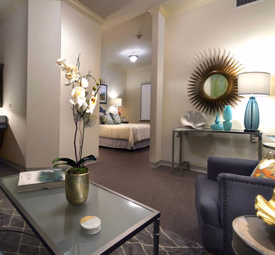 Open-plan living room and bedroom suite with a glass coffee table and potted orchid in the foreground, a seating area to the right, and a bed and console table with lamp and sunburst mirror in the background.