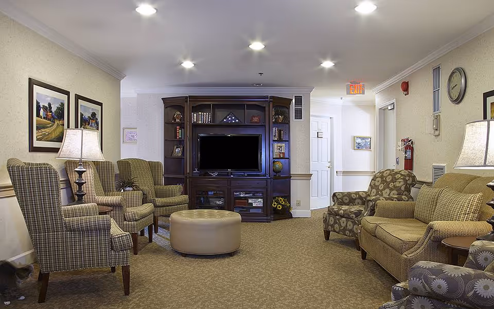 A cozy living room area in a senior living facility with patterned armchairs and a sofa arranged around a central ottoman. A large wooden entertainment center with a flat-screen TV and decorative items is against the back wall. The room is softly lit with table lamps and recessed ceiling lights, and framed landscape paintings hang on the walls.