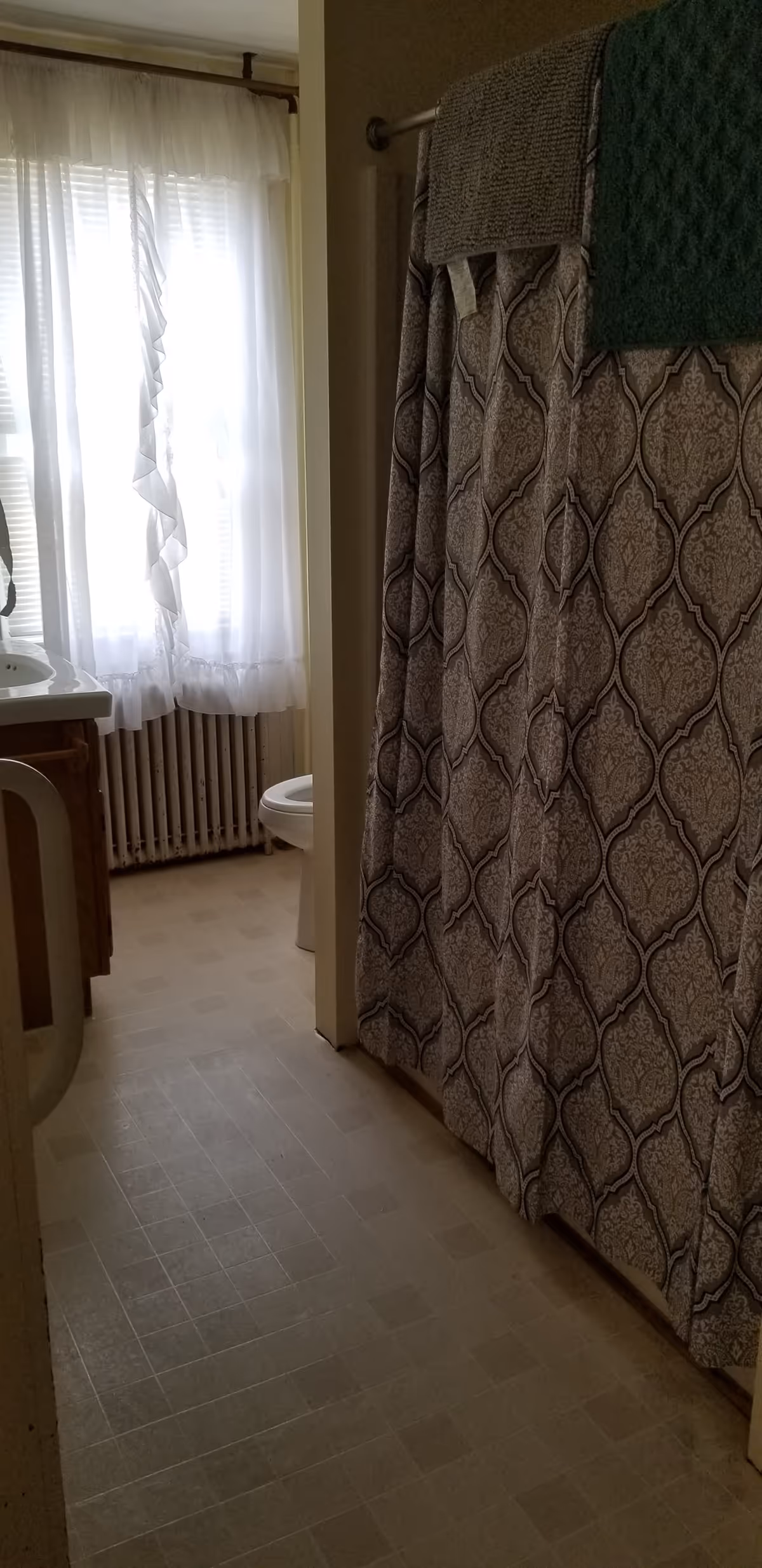 Narrow bathroom with a patterned shower curtain and towels, a sink and toilet next to a window with sheer curtains and a radiator.