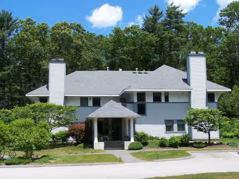 Exterior view of a two-story senior living facility with a gray roof and surrounding trees.