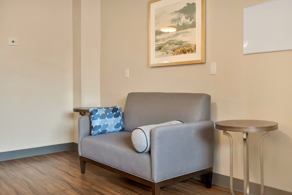 A small gray loveseat with two decorative pillows, one blue patterned and one cylindrical gray, placed against a beige wall. There are two small round side tables on either side of the loveseat. A framed landscape painting hangs on the wall above the loveseat.