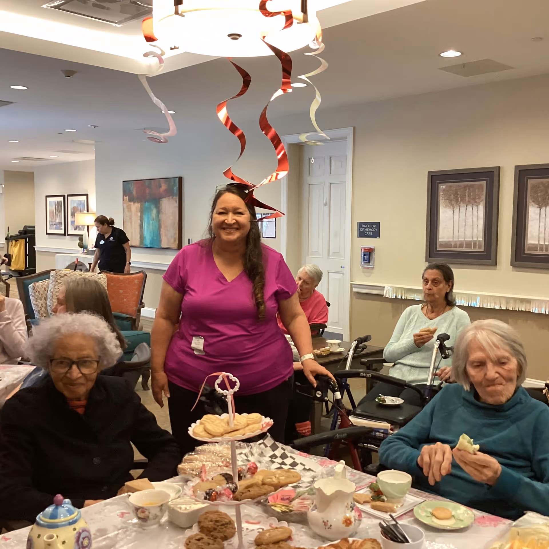 A smiling staff member stands with elderly residents seated around a table laid out with tea, cookies, and pastries in a decorated communal dining/activity room.