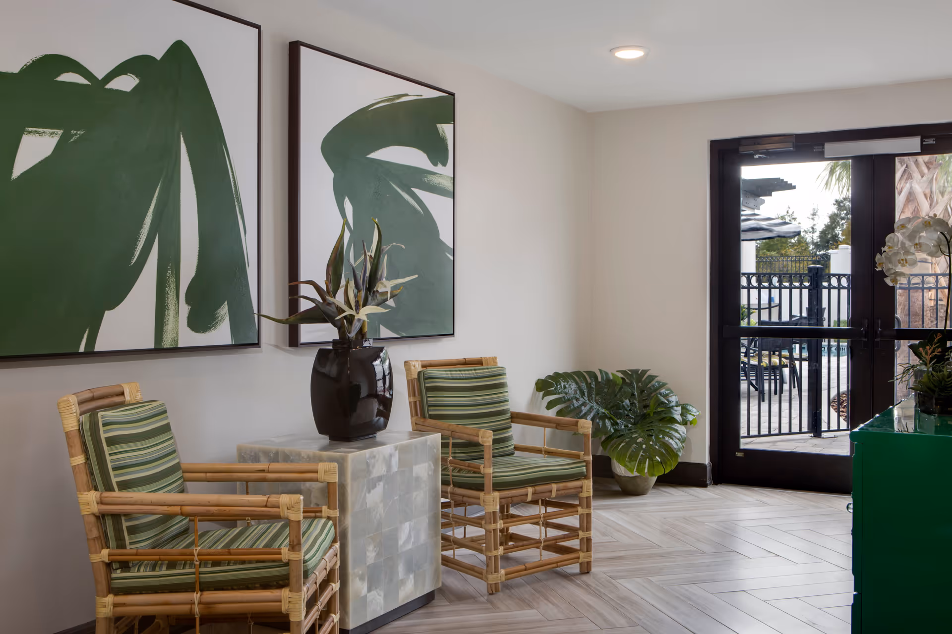 Small seating area with two striped-cushioned bamboo chairs, a side table with a vase, large green abstract wall art, and glass doors leading outside.