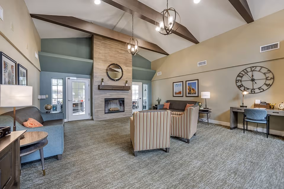 A spacious living room with vaulted ceilings and exposed wooden beams. The room features a stone fireplace with a round mirror above it, two striped armchairs, two gray sofas with orange pillows, a desk with a chair and a large decorative wall clock, and several framed pictures on the walls. There are two glass doors on either side of the fireplace, and the room is well-lit with hanging pendant lights and table lamps.