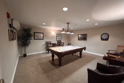 A recreation room with a pool table in the center, a dartboard on the wall, several chairs arranged around the room, framed pictures on the walls, a potted plant in the corner, and overhead lighting fixtures.