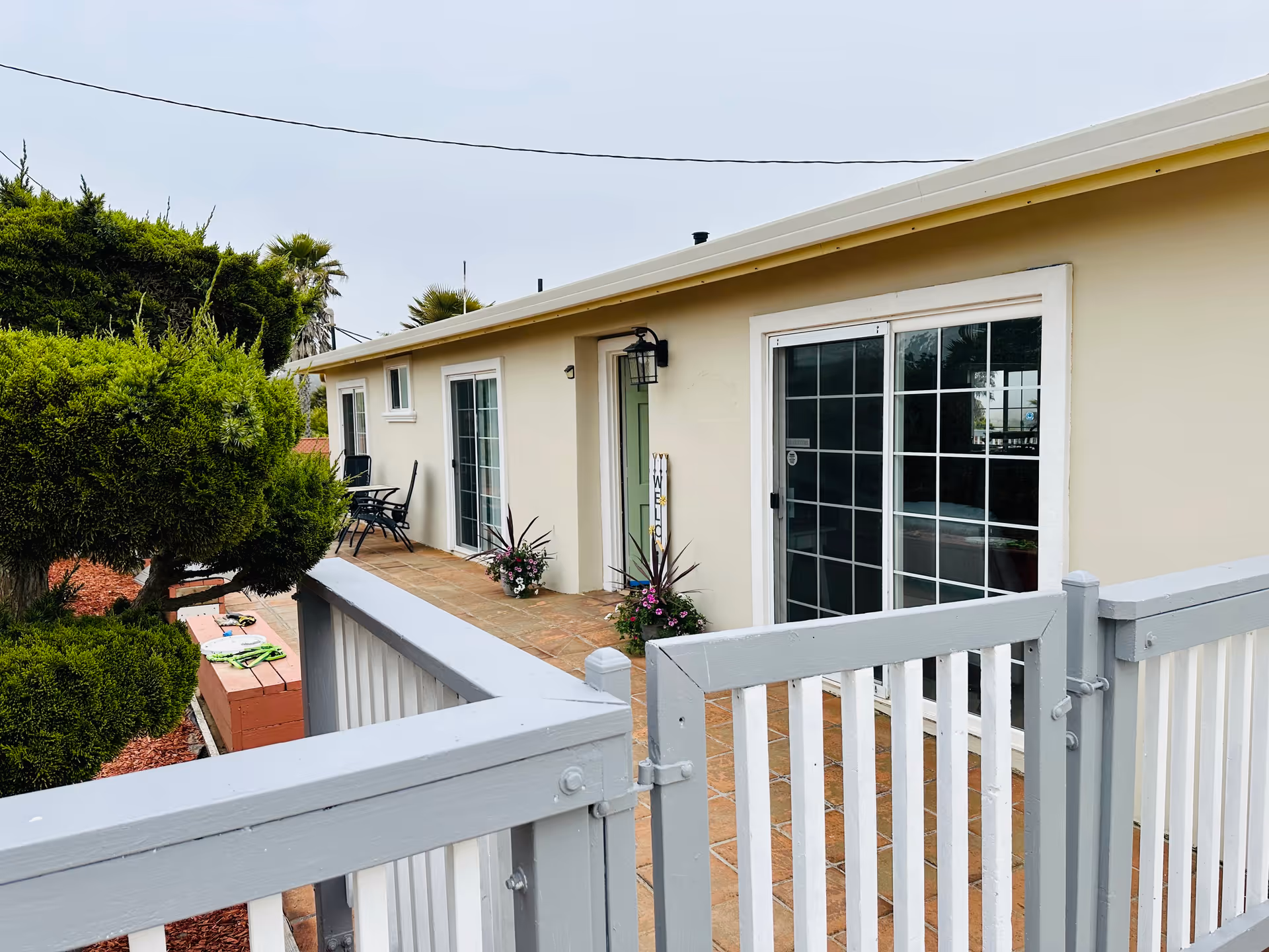 Outdoor patio area of a single-story building with sliding glass doors and a green entrance door. There are potted plants and a welcome sign near the door. The patio is enclosed by a white and gray wooden fence, and there are trimmed bushes and trees nearby.