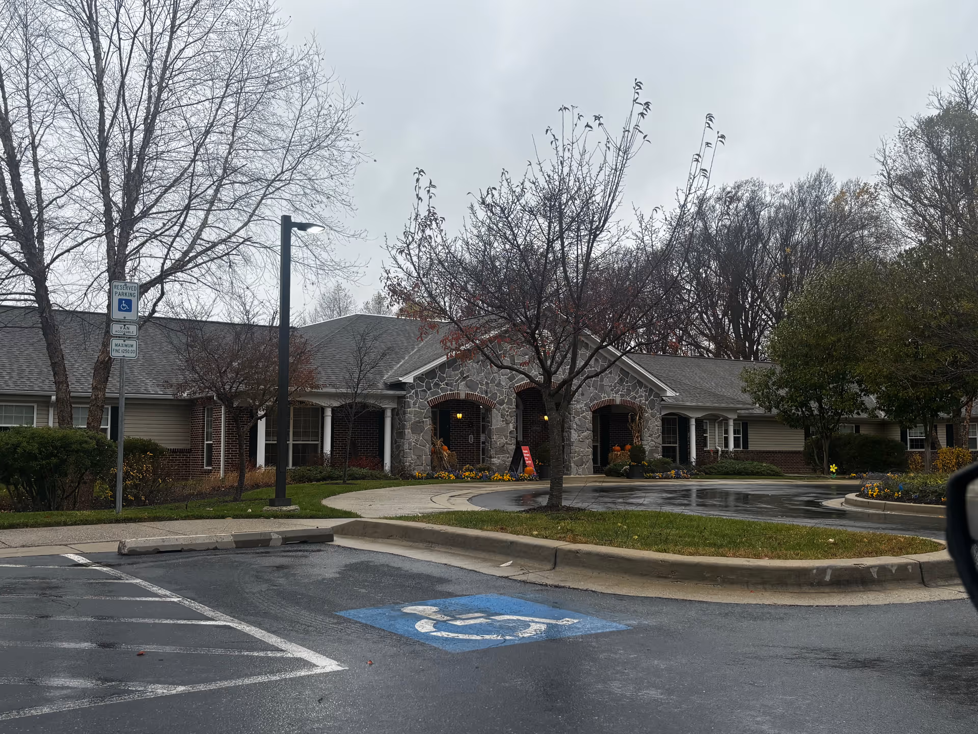 Exterior view of a single-story building with stone and brick facade, surrounded by leafless trees and a wet parking lot with a handicapped parking space in front. The building has a covered entrance with columns and some fall decorations near the door.