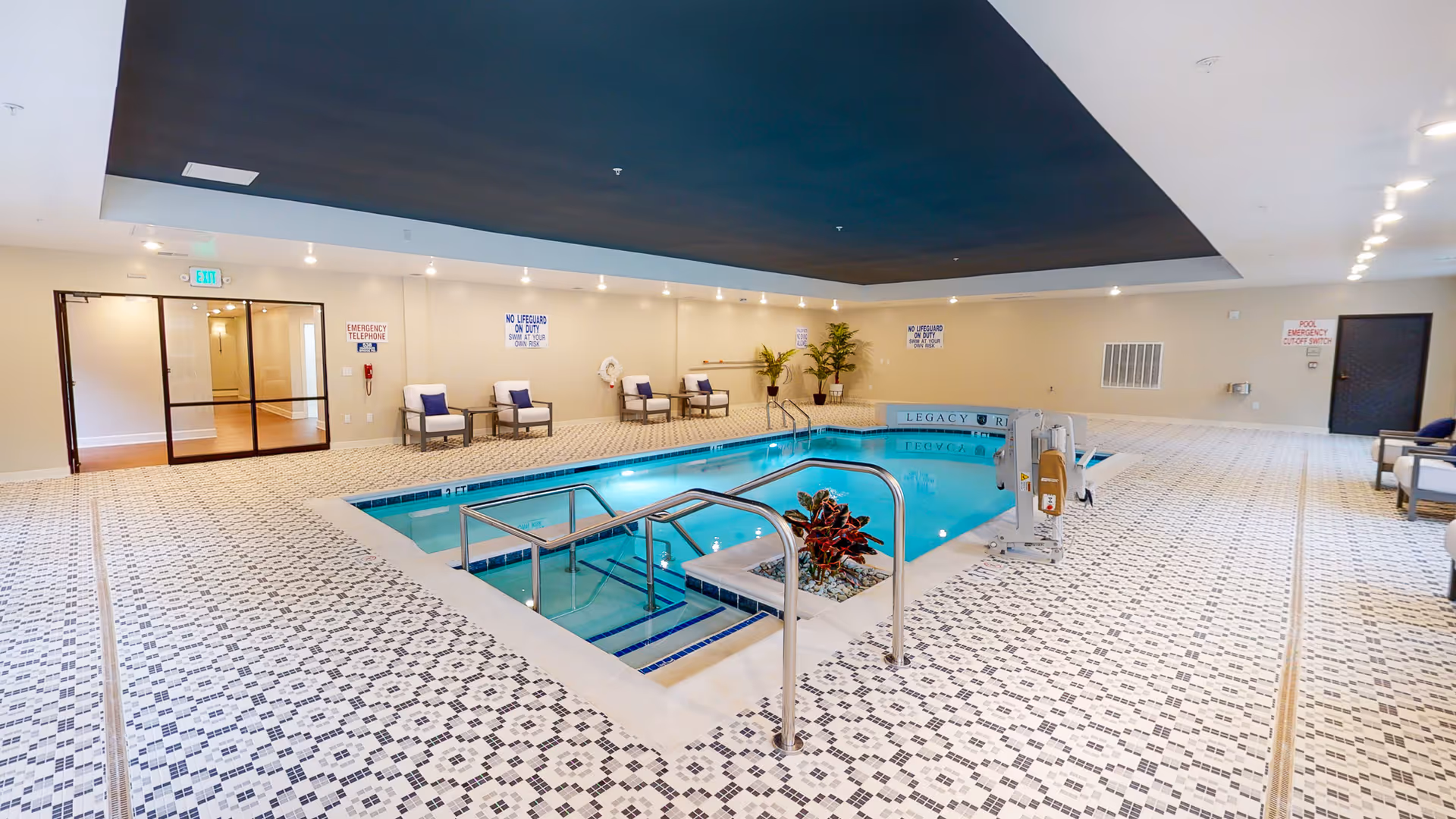 Indoor swimming pool area with patterned tile flooring, several chairs along the walls, handrails leading into the pool, and a pool lift for accessibility. The ceiling above the pool is dark, contrasting with the light-colored walls and floor. There are emergency and safety signs on the walls.