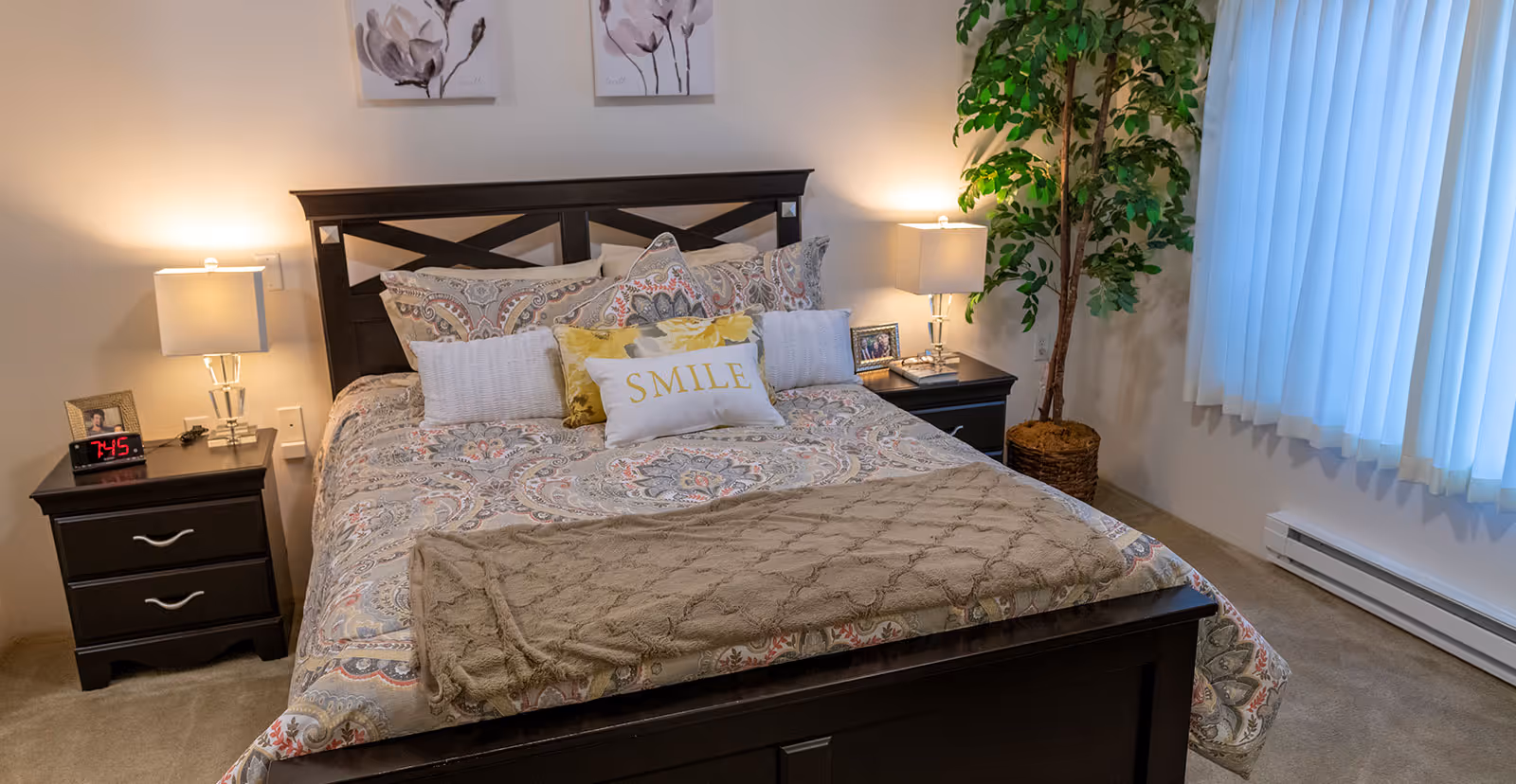 Sunlit bedroom featuring a neatly made dark-wood bed with patterned bedding and a 'SMILE' pillow, flanked by nightstands, lamps, and a potted plant by a window.