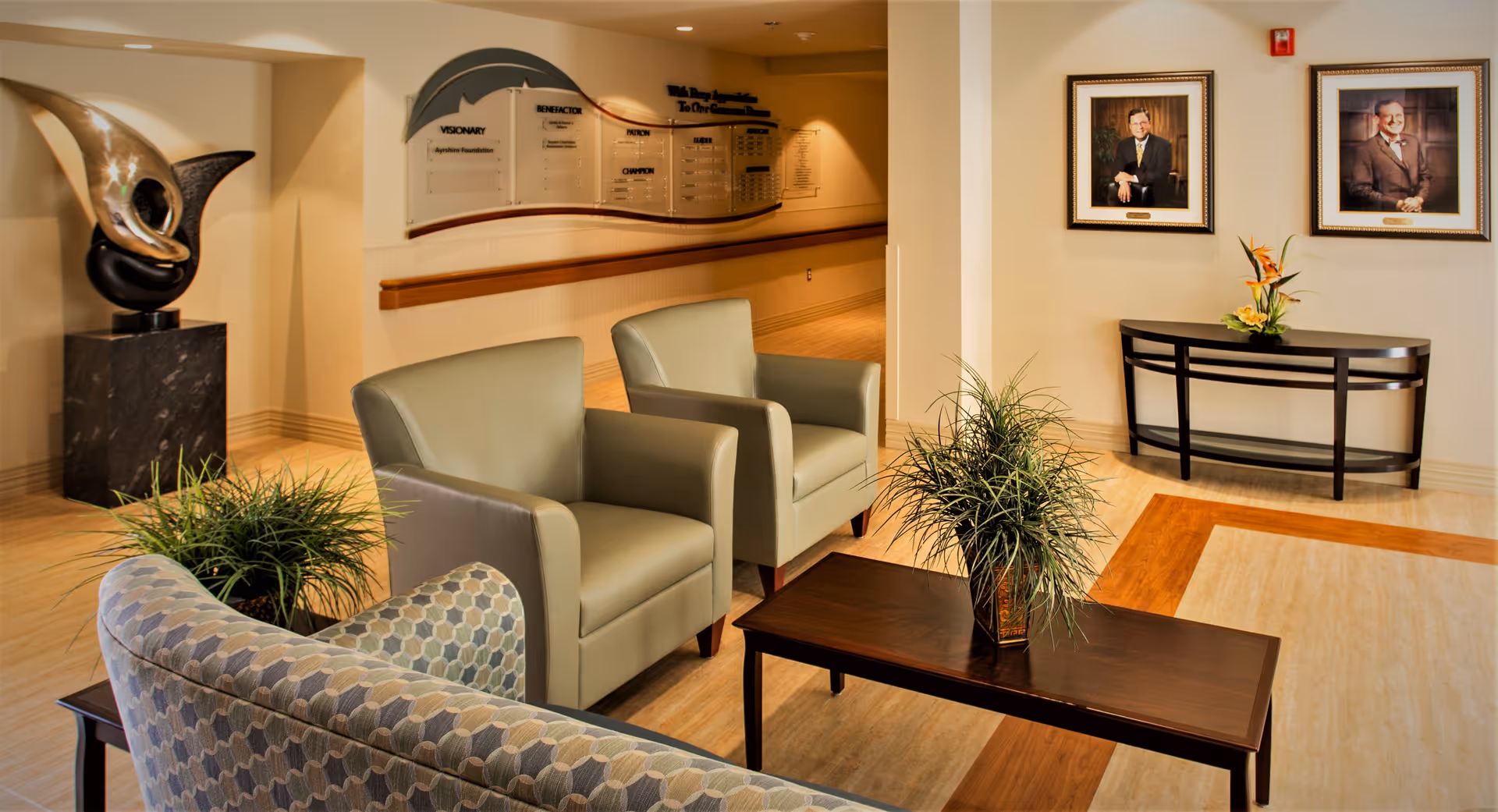 A well-lit seating area in a senior living facility with two beige armchairs, a patterned sofa, a dark wooden coffee table with a potted plant, and a console table with a flower arrangement. The walls feature two framed portraits and a large decorative plaque. There is also a modern abstract sculpture on a pedestal in the corner.