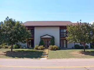 Two-story senior living facility exterior with central entrance, steps, and surrounding trees.