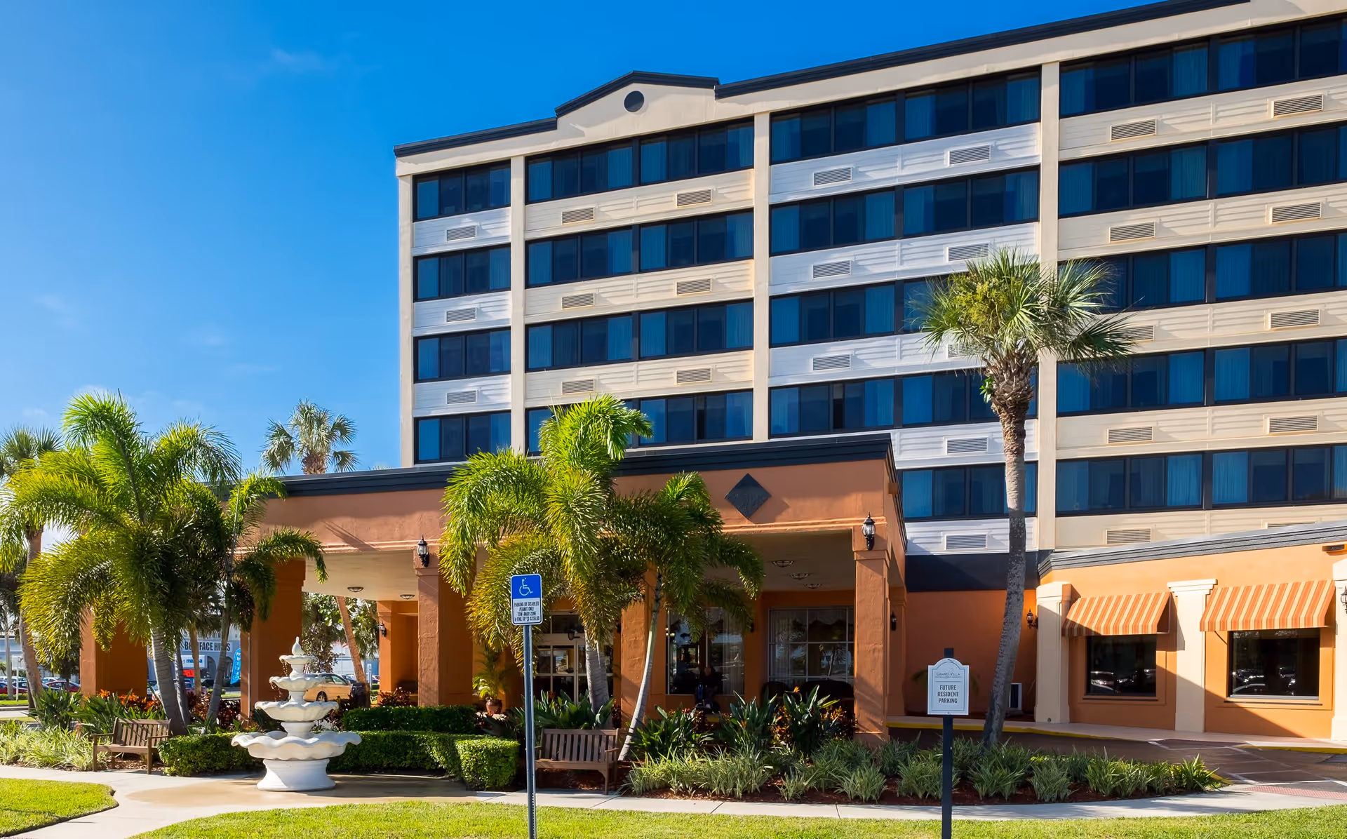Exterior view of a multi-story senior living facility with a covered entrance, palm trees, a white tiered fountain, benches, and well-maintained landscaping under a clear blue sky.