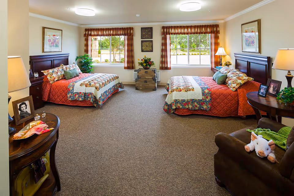 A cozy bedroom in a memory care community featuring two twin beds with floral and patchwork quilts, wooden headboards, and matching bedside tables with lamps. The room has two large windows with plaid curtains letting in natural light, framed artwork on the walls, a small chest between the windows with a flower arrangement, and a comfortable armchair with a stuffed pig toy and green blanket.